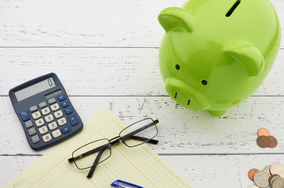 A piggy bank beside a calculator, notepad, reading glasses, and coins on a white desk